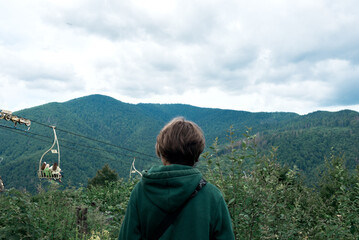 Man standing in mountain top, looking on mountain landscape with pine trees, cloudy sky and cableway, back view. Carpathian mountains, Ukraine