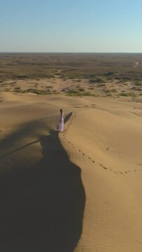 Young lonely woman with long hairs in white dress staying on the top of sand beige dune in desert. Drone is flying around. Aerial view. Vertical Video