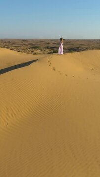 Young lonely woman with long hairs in white dress staying on the top of sand beige dune in desert. Drone is approaching. Aerial view. Vertical Video
