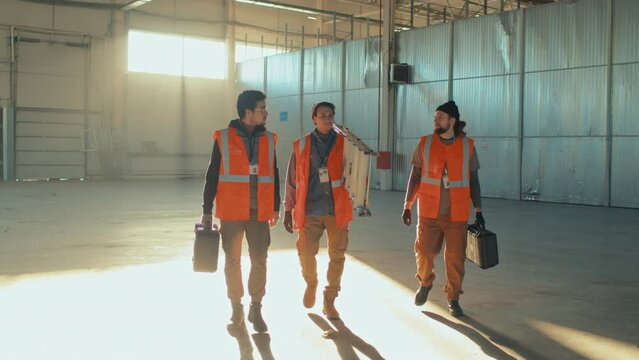 Full length tracking footage of three diverse male construction workers walking along spacious building with tool boxes and ladder and speaking