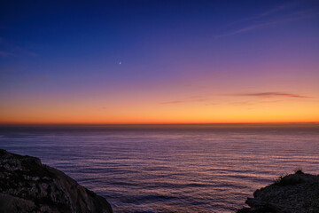 Sunset over sea waters, dark cliffs and crescent moon in the sky.