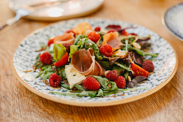 Close up of served fresh salad with jamon, raspberries, tomatoes cherry, cheese and mixed lettuce on plate, dressed olive oil, balsamic vinegar and spices on wooden table at restaurant. Food at event.