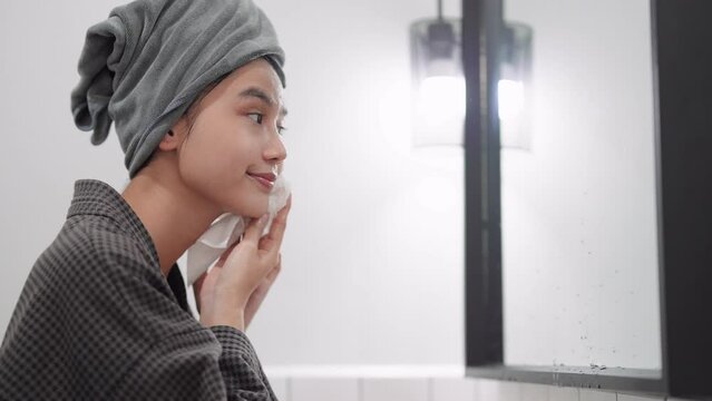 An Asian woman is patting her face dry after cleansing, gently drying her face with a towel in front of the bathroom mirror. Capture this skincare routine for a refreshing image