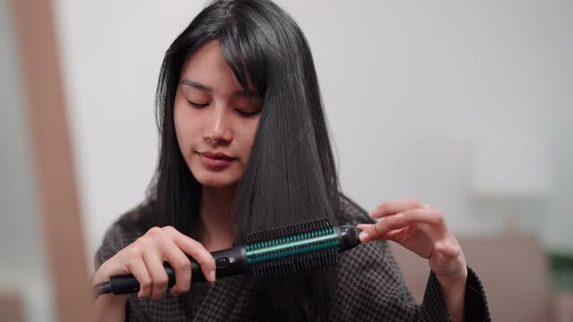 Asian woman using a hair straightener to achieve smooth hair in front of a large mirror after washing and styling at home. Showcase the beauty routine for enhanced sales
