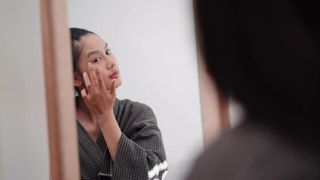 Asian woman applying concealer on her face in front of a large mirror after washing and styling her hair at home. Capture her beauty routine.