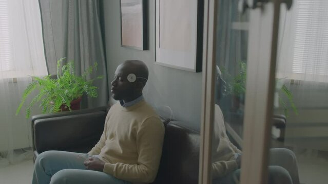 Black man with electrodes on head sitting with eyes closed on sofa during EEG test in sleep clinic
