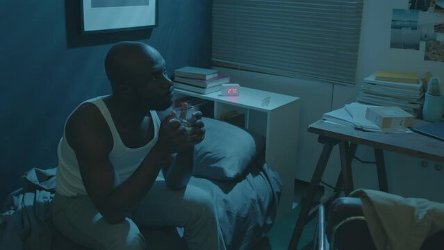High angle shot of sleepless Black man sitting on bed in dark room and drinking herbal tea while suffering from insomnia at night
