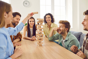 Obraz premium Portrait of excited happy young friends guys and girls playing together with wooden building blocks at home sitting at the table enjoying time together. Home leisure and board games concept.