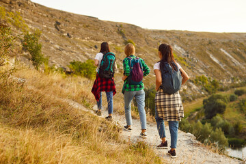 Back view of a group of young girls tourists with backpacks hiking in nature walking in a row. Active female friends trekking outdoors. Adventure, vacation travel and tourism concept.