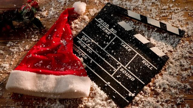 Snow covers a movie clapper board, a Christmas hat, and a spruce branch on a wooden table. A concept based on the release of Christmas holiday movies and film productions.