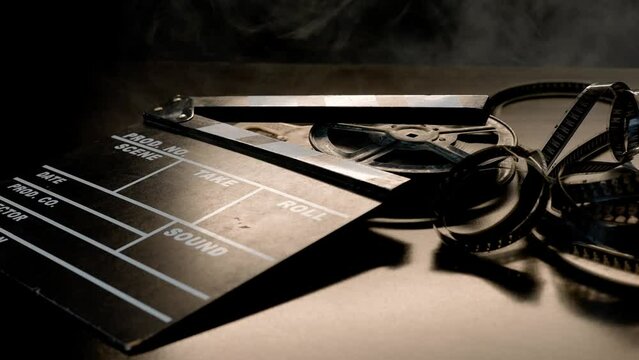 A clapper board and a reel of film roll on a wooden table in puffs of smoke. Film industry equipment in studio on black background.