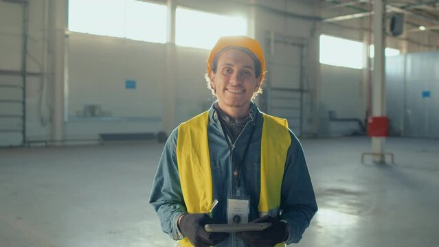 Medium portrait of smiling male Biracial builder in safety hardhat and workwear with tablet posing at looking at camera during workday in empty indoor construction site