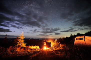 Romantic couple enjoying at starry sky. Young people sitting in chairs and warming by campfire near tourist tent. Travelling couple having a rest together. Night camping. - obrazy, fototapety, plakaty
