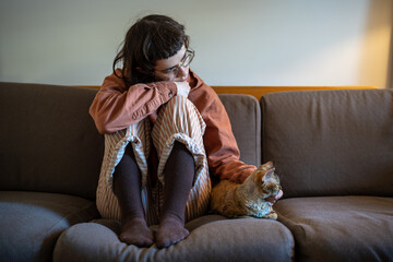 Upset sad depressed teenager sitting on sofa, putting head on knees, stroking beloved breed cat...