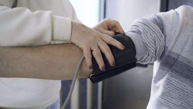 An elderly woman measures her husband's blood pressure. She puts the cuff on her arm.