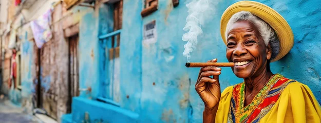 Photo sur Plexiglas Havana Elderly Woman Enjoying a Cigar Against a Blue Wall in Havana. Smiling Senior Lady in Yellow Dress and Straw Hat with Cigar in Cuba
