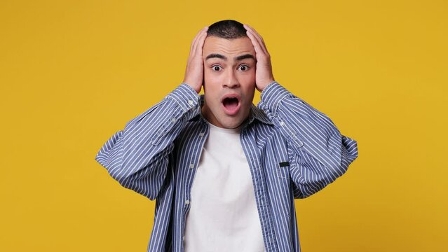 Young scared indignant astonished sad shocked middle eastern man he wear blue shirt white t-shirt look camera hold head say what isolated on plain yellow background studio portrait. Lifestyle concept