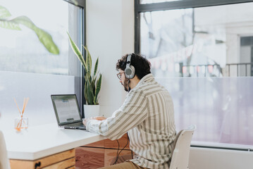 Male employee sitting on working desk next to the window is listening to music on the headphones and working on his daily tasks