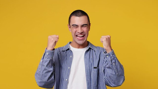Excited jubilant overjoyed happy young middle eastern man he wear blue shirt white t-shirt doing winner gesture celebrate clenching fists say yes isolated on plain yellow background. Lifestyle concept