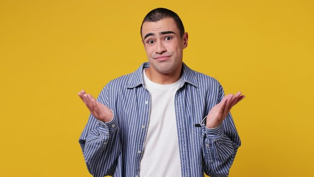 Hesitant indecisive fun shy young middle eastern man wear blue shirt white t-shirt feels doubtful spreading hands say oops ouch oh omg i am so sorry isolated on plain yellow background studio portrait