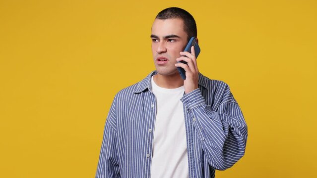 Angry sad tired young middle eastern man he wear blue shirt white t-shirt hold use talk on mobile cell phone do not want listen ask what bad fake news scream swear isolated on plain yellow background