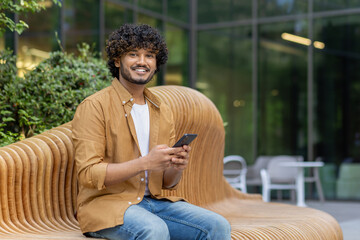 Portrait of a young Indian man waiting for an appointment sitting on a bench on the street, resting, holding a phone and smiling at the camera