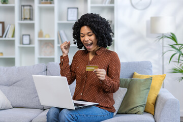 African american young woman happy with success looking at laptop sitting on sofa at home and...