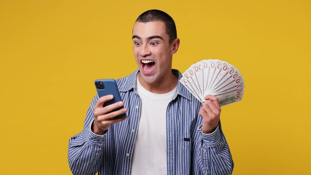 Excited happy joyful young middle eastern man he wear blue shirt white t-shirt use mobile cell phone hold win fan of cash money in dollar banknotes isolated on plain yellow background studio portrait