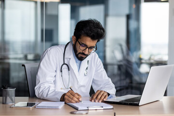 Concentrated young Indian male doctor in white coat sitting in office at table with laptop and writing medical results and documents