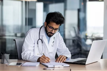 Poster Concentrated young Indian male doctor in white coat sitting in office at table with laptop and writing medical results and documents © Tetiana