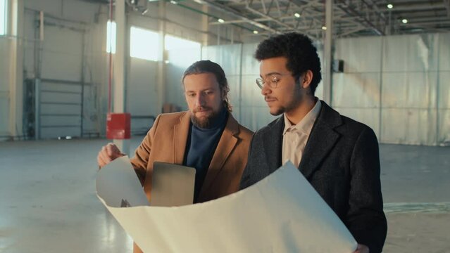 Medium side shot of Caucasian and Biracial business associates looking at building plan and discussing new project implementation standing in empty spacious room