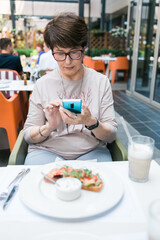 European mature woman sitting at table on terrace of restaurant drinking milk cocktail and chatting on mobile app