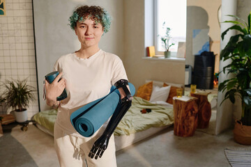 Portrait of positive teenage girl with prosthetic arm holding yoga mat and water bottle
