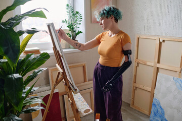 Pensive girl with prosthetic arm focused on painting picture on canvas