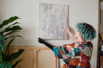 Artist with prosthetic arm hanging picture on wall in her apartment