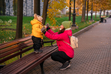 Cute baby toddler child in a yellow jacket in an autumn park stands on a bench with a young mother