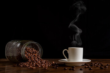Cup of steaming coffee next to an overturned jar with coffee beans on a wooden table with more loose beans, front view and close-up.
