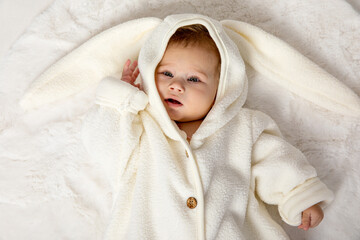 Adorable funny newborn baby girl wearing a white warm bunny snow suit on a white background. Beautiful baby girl in a warm winter white suit lying on a bed. Toys and goods for children. Top view