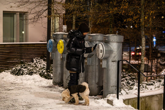 Stockholm, Sweden, A young woman throws garbage in a shute with a dog on a leash in a resdiential complex at night in the winter.
