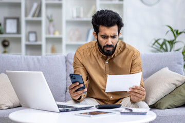 Worried young Indian man sitting on sofa at home, working on laptop, looking sadly at documents and bills, holding phone.