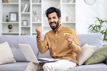 Portrait of a happy young man sitting on the sofa at home with a laptop, holding a credit card and looking at the camera with a victory gesture. - Powered by Adobe