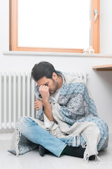 Man freezing at home and sitting by the radiator. Freezing guy warming close to heating system. High prices for gas and other Communal expenses