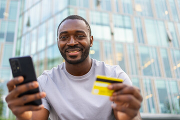 African American man shopping online holding smartphone paying with gold credit card Guy on urban...