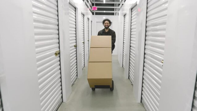 Happy bearded male African American pushing trolley cart with large carton boxes in front of him along white corridor in a climate-controlled storage. High quality 4k footage