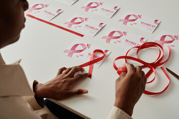 Hands of young woman with pink ribbon making symbols of breast cancer and fixing them on handmade...