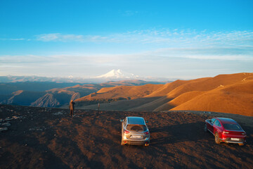 Panoramic picturesque view of snow-covered Elbrus in the early morning from the mountain plateau with dusty cars on the top. Shooting from a drone.