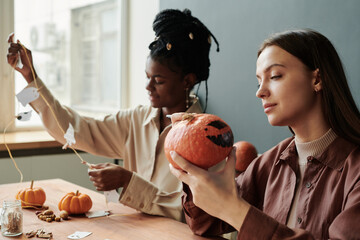 Young brunette woman looking at spooky Halloween pumpkin in her hands while sitting against female friend creating decorations