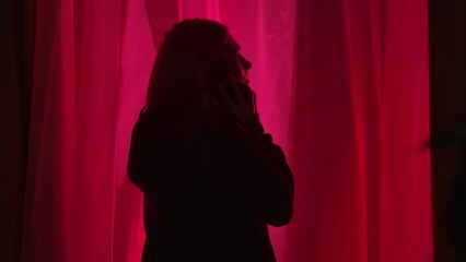 Back view of a woman standing near a window in a dark room illuminated by police lights, close up. The anxious woman is talking on her smartphone, grabbing her head with worry and hopelessness.