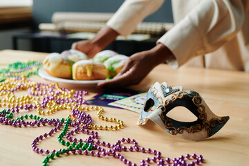 Carnival mask and colorful beads lying on wooden table while young woman putting plate with...