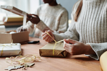 Hands of young unrecognizable woman in sweater tying ribbon around giftbox wrapped into craft paper...